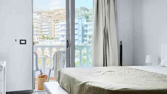 Utsikt från ett hotellrum, med säng och en öppen balkongdörr. Balkong med stolar och utsikt över byggnader. (AI-genererad text)