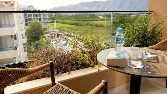 En balkong med utsikt över grönska och berg, med en bord innehållande böcker, glas, och en vattenflaska. (AI-genererad text)