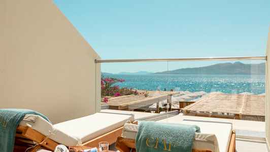 En solstol med en grön handduk vid en balkong med glasräcke, med utsikt över havet och en strand med parasoller. (AI-genererad text)