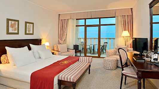 Ett hotellrum med dubbelsäng, sittgrupp och balkong med havsutsikt. (AI-genererad text)