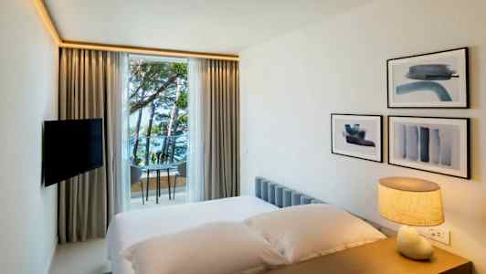 Ett hotellrum med en säng, en lampa på bordet, väggdekorationer och ett fönster som vetter mot havet och träd. (AI-genererad text)