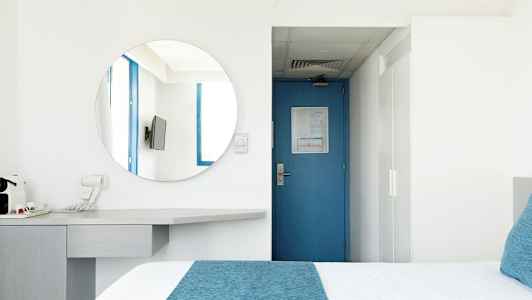 Ett hotellrum med en blå dörr, en rund spegel, en TV och en säng med blå detaljer. (AI-genererad text)