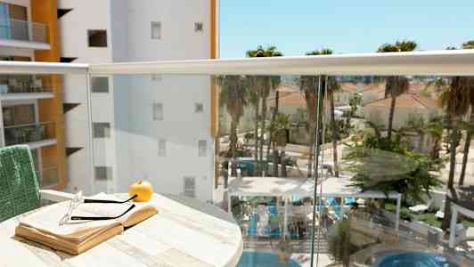 En balkong med ett öppet bok, ett par glasögon och ett äpple på ett bord. I bakgrunden syns en pool med omgivande palmer och byggnader. (AI-genererad text)