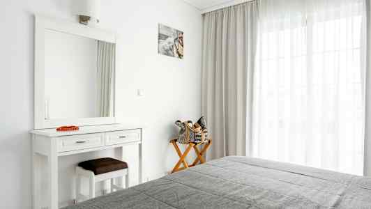 Ett ljust sovrum med en spegel, ett bord, och en stol vid fönster med gardiner. (AI-genererad text)