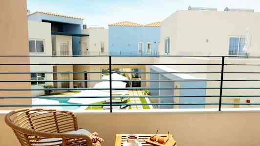 Utsikt från en balkong mot en innergård med en liten pool, omgiven av ljusa byggnader. På balkongen finns en sittstol och ett bord med glasögon och en kaffekopp. (AI-genererad text)