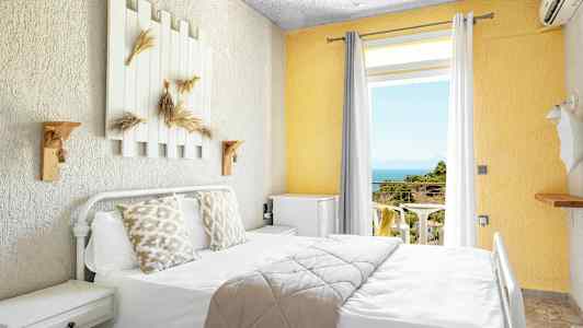 Ljust hotellrum med säng, dekorerade kuddar och utsikt mot balkong med hav och grönska. (AI-genererad text)