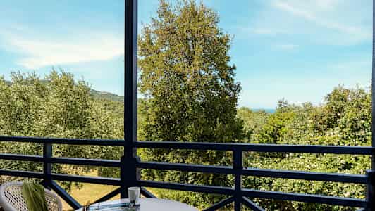 Utsikt från en balkong med bord och stolar mot träd och hav i bakgrunden. (AI-genererad text)