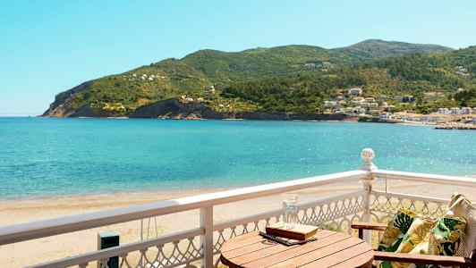 Utsikt från en balkong mot havet och grönskande kullar, med en trästol och ett bord. (AI-genererad text)