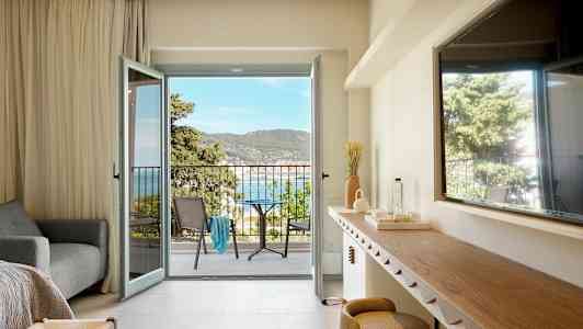 Ett hotellrum med öppen balkongdörr som visar utsikt över havet och omgivande grönska. (AI-genererad text)