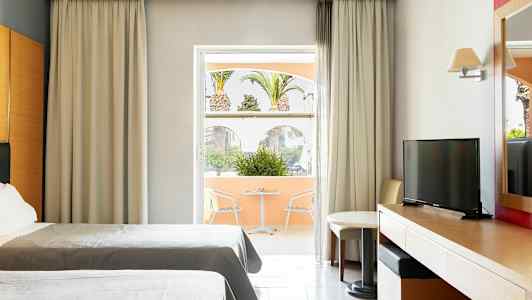 Hotellrum med två sängar, skrivbord, TV och en sittgrupp vid ett fönster ut mot en terrass med utsikt över palmer. (AI-genererad text)