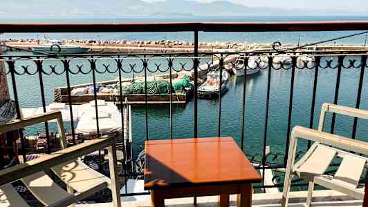 Utsikt från en balkong med bord och stolar mot en hamn med båtar och ett stenpir. (AI-genererad text)