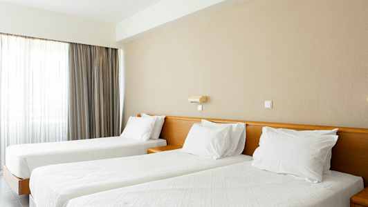 Två bäddar med vita täcken och kuddar i ett ljust rum med gardiner. (AI-genererad text)