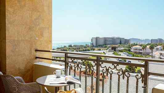 En balkong med ett runt bord, två stolar och utsikt mot byggnader, vägar och havet. (AI-genererad text)