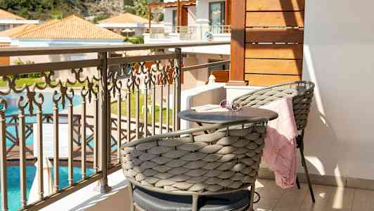 En balkong med två sittplatser, ett litet bord, och en fin utsikt över en swimmingpool och byggnader. (AI-genererad text)