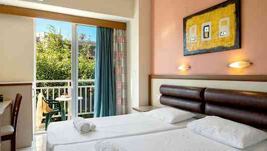 Ett hotellrum med dubbelsäng, sänglampor, skrivbord, och en balkong med utsikt mot grönska. (AI-genererad text)