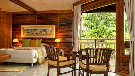 Ett träbeklätt sovrum med en dubbelsäng, två stolar och ett bord framför en stor öppning mot en balkong med trädgårdsutsikt. (AI-genererad text)