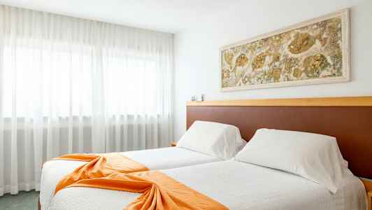 Ett hotellrum med två sängar klädda i vita sängkläder och orange prydnadssjalar, en väggdekoration ovanför huvudgavlarna samt gardiner framför ett stort fönster. (AI-genererad text)