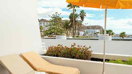 Solstol med handduk under parasoll på en balkong med utsikt mot palmer och byggnader. (AI-genererad text)