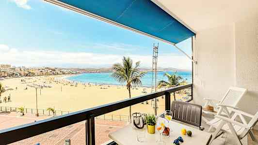 Utsikt från en balkong mot en sandstrand och hav, med bord och stolar i förgrunden. (AI-genererad text)