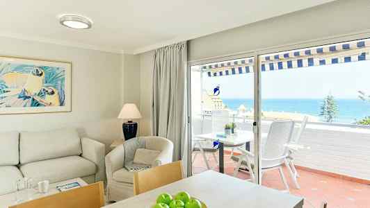 Interiör av ett vardagsrum med soffa, fåtölj och bord, och utsikt över havet genom glasdörrar till en terrass. (AI-genererad text)