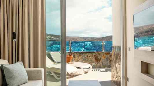 En utsikt från ett rum mot en terrass med solstolar och glasräcke, landskap och himmel synliga i bakgrunden. (AI-genererad text)