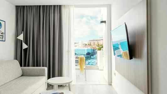 Ett vardagsrum med soffa, bord, tv och en öppen balkongdörr som visar utsikt över byggnader och hav. (AI-genererad text)