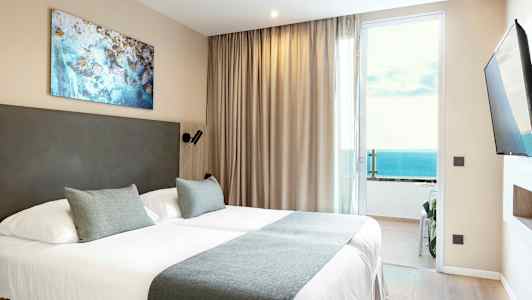 Ett hotellrum med en dubbelsäng, beige gardiner och havsutsikt genom en glasdörr. (AI-genererad text)