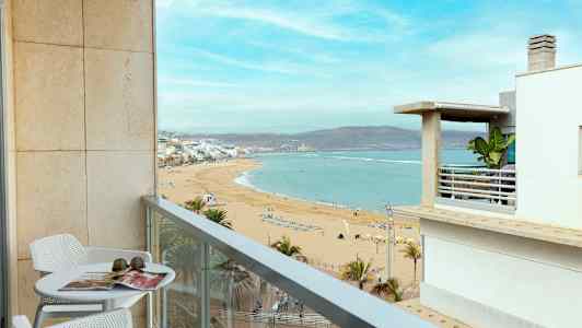 En balkong med bord och stolar erbjuder utsikt över en sandstrand och havet. (AI-genererad text)