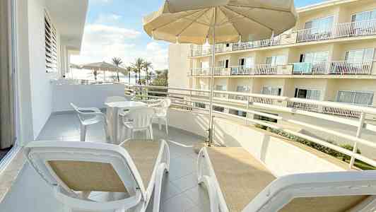 Balkong med solstolar under ett parasoll och utsikt över byggnader och palmer. (AI-genererad text)