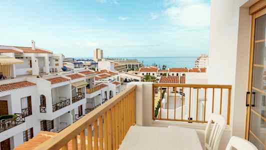 Utsikt från en balkong med ett bord och stolar över vita byggnader med röda tak och havet i bakgrunden. (AI-genererad text)