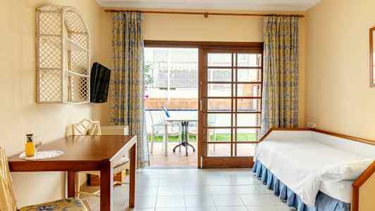 Ett hotellrum med en enkelsäng, bord och stolar, samt utsikt mot en terrass med möbler. (AI-genererad text)