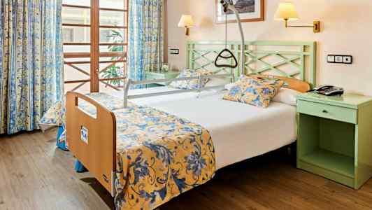 Hotellrum med en säng utrustad med stödhandtag och gul- och blommönstrade sängkläder. (AI-genererad text)