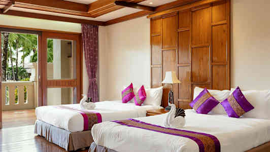 Två bäddar med vita lakan och dekorativa kuddar i ett rum med trädetaljer och en balkong med utsikt över träd. (AI-genererad text)