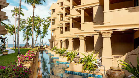 Byggnad med flera balkonger, omgivna av gräsmattor, palmer och en pool. (AI-genererad text)