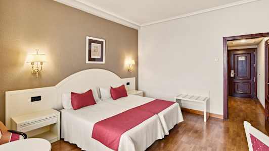 Ett hotellrum med en dubbelsäng som har röda detaljer, vägglampor och en dörr i bakgrunden. (AI-genererad text)