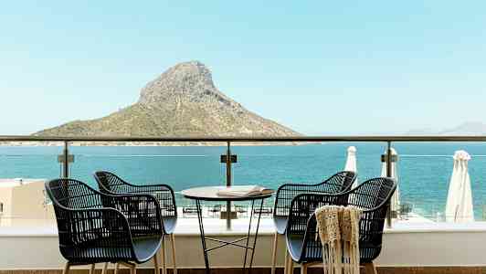 Fyra stolar och ett bord på en terrass med utsikt över havet och en bergstopp. (AI-genererad text)