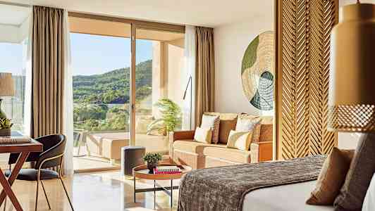 Ett hotellrum med en säng, sittgrupp och utsikt över grön natur genom glasdörrar. (AI-genererad text)