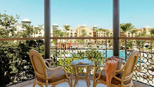 Utsikt från en balkong med två rottingstolar och ett bord, mot en pool och hotell omgivna av palmer. (AI-genererad text)