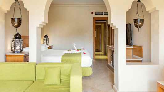 Ett hotellrum med en säng dekorerad med en formad handduk och blommor, en grön soffa i förgrunden och utsmyckade lampor. (AI-genererad text)