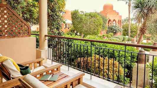 En terrass med trästolar och utsikt över en lummig trädgård och byggnader med bågformade fönster. (AI-genererad text)