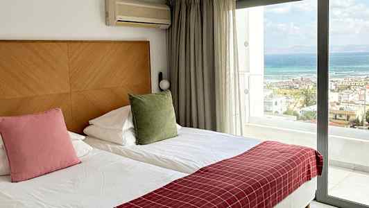 Ett hotellrum med två enkelsängar, en stor glasdörr som visar utsikt mot havet och bekvämligheter som luftkonditionering. (AI-genererad text)