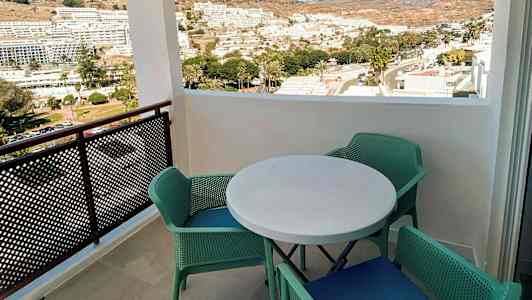 Utsikt från en balkong med ett runt bord och fyra gröna stolar, med utsikt över en stad och berg i bakgrunden. (AI-genererad text)