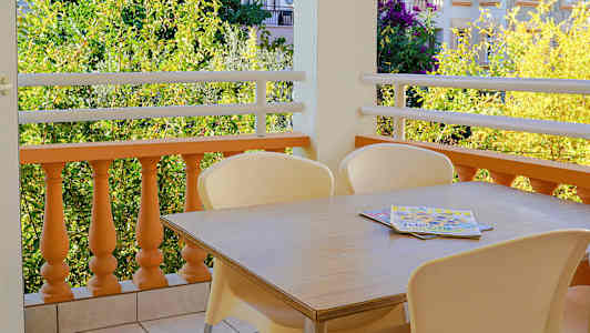 Bild av en balkong med ett bord och stolar på klinkerbelagt golv samt utsikt över grönska och byggnader. (AI-genererad text)