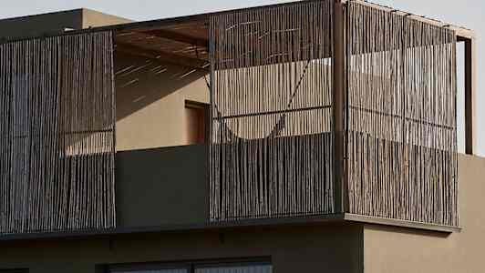 En modern byggnad med en balkong inkapslad med trästavar och glasfönster. Framför syns vegetation i ett ökenlandskap. (AI-genererad text)