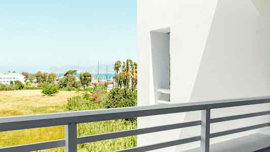 Utsikt från en balkong med bord och stol, vita byggnader och grönska i bakgrunden. (AI-genererad text)