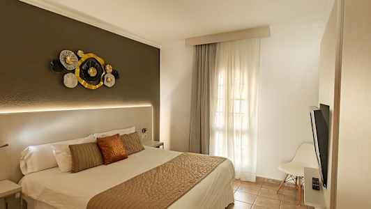 Ett hotellrum med en dubbelsäng, dekorativ vägghängd konst ovanför sänggaveln och ett fönster med ljusa gardiner. (AI-genererad text)