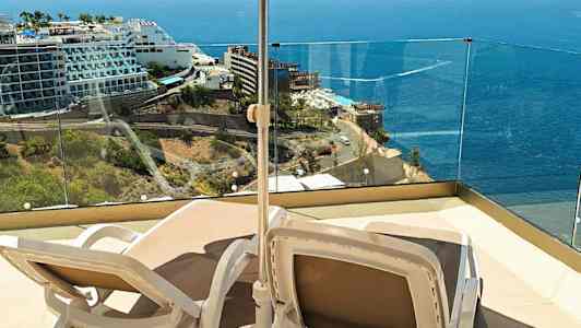 Två solstolar under ett parasoll på en terrass med utsikt över havet och närliggande hotell. (AI-genererad text)