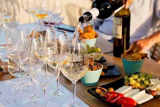 Ett bord med glas fyllda med vitt vin som serveras, omgivna av olika tilltugg som ost, bröd och frukt. (AI-genererad text)