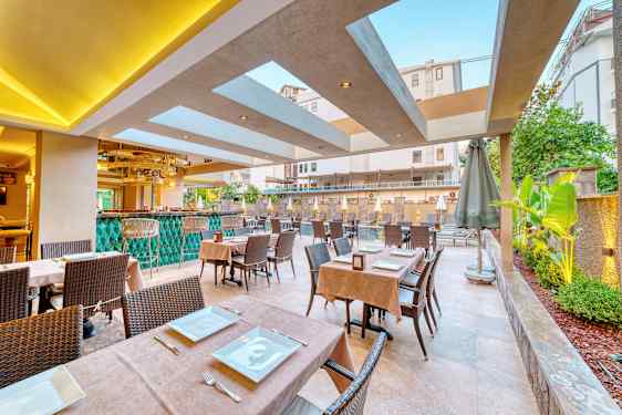 Utomhusrestaurang med bord dukade med tallrikar och bestick, samt gröna växter och pool i bakgrunden. (AI-genererad text)