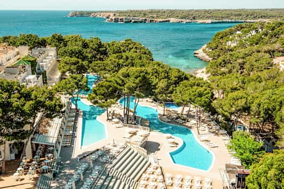 En vy över en semesteranläggning med pooler omgivna av solstolar och parasoller, belägen intill ett turkost hav med frodig vegetation i bakgrunden. (AI-genererad text)